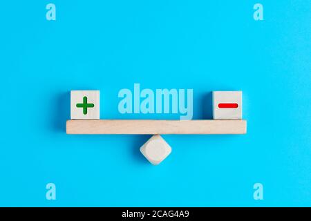 Plus and minus or positive and negative symbols on wooden blocks are in balance on a wooden seesaw. Blue background, flat lay view. Pros and cons equi Stock Photo