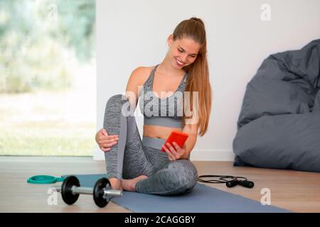 young fit woman using smartphone during the workout - female sporty girl at home sitting on yoga mat looking to the mobile phone Stock Photo