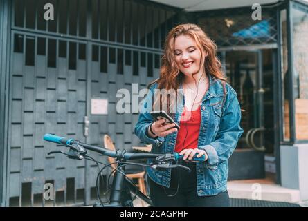 Portrait of Red curled long hair caucasian teen girl on the city street walking with bicycle using the smartphone with earphones. Natural people beaut Stock Photo