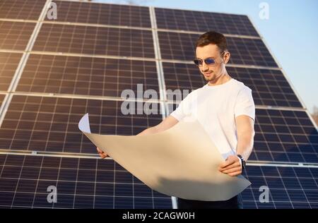Young businessman is looking into a white paper plan standing by a big solar panel, sun is shining brightly on the face, eco-friendly concept Stock Photo