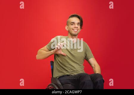 Shows tiny. Caucasian young disabled man's portrait isolated on red studio background. Beautiful male model in casual style. Concept of human emotions, facial expression, sales, ad, inclusion. Copyspace. Stock Photo