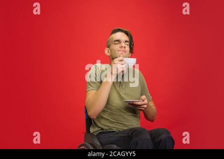 Joyful coffee, tea time. Caucasian young disabled man's portrait on red studio background. Beautiful model in casual style. Concept of human emotions, facial expression, sales, ad, inclusion. Copyspace. Stock Photo