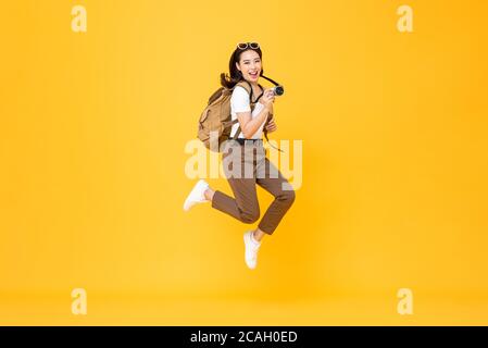 Young pretty Asian woman tourist backpacker smiling and jumping with camera in hand isolated on yellow background Stock Photo