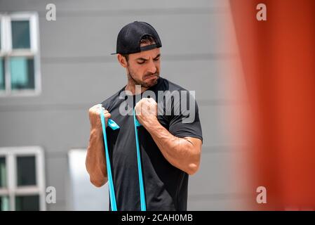 Fit muscular Latino sports man doing bicep curl exercise with resistance band outdoors at home in sunlight Stock Photo