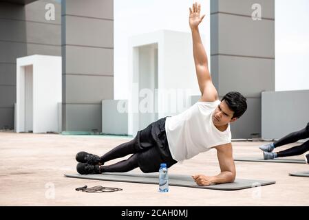 Athletic Indian man doing side plank exercise outdoors on rooftop with friends Stock Photo