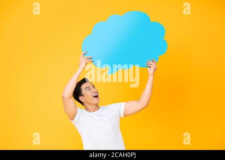 Excited smiling young handsome Asian man holding and looking up to speech bubble with empty space for text on yellow background Stock Photo