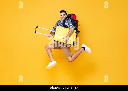 Young excited Caucasian male tourist with baggage jumping in mid-air ready to travel isolated on colorful studio yellow background Stock Photo