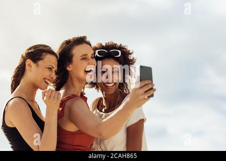 Portrait of three women friends enjoying a selfie standing outdoors. Happy woman taking selfie with friends using mobile phone. Stock Photo