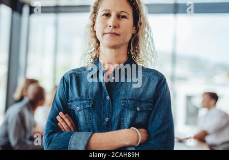 Portrait of a confident female business professional standing with her arms crossed. Businesswoman in casuals with colleagues meeting at the back.