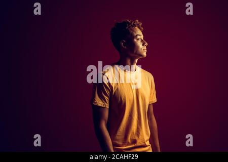 Freestyle. Mulatto guy standing isolated on dark red in yellow light looking aside pensive Stock Photo