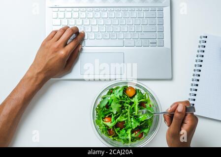 top view of the hands of a black woman using a laptop while eating a salad on the top of a white desk Stock Photo