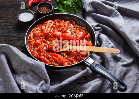 close-up of ragout with whole plum tomatoes, onion slices, ground italian pork sausages and spices in a skillet on a dark oak wooden table with grey c Stock Photo