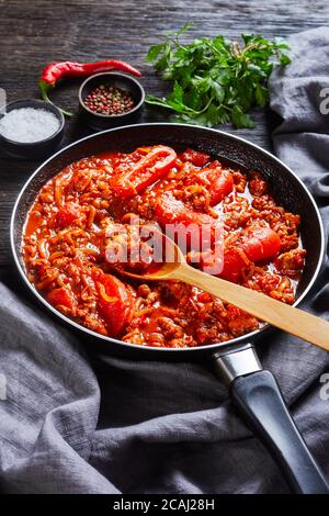 ragout with whole plum tomatoes, onion slices, ground italian pork sausages and spices in a skillet on a dark oak wooden table with grey cloth, vertic Stock Photo
