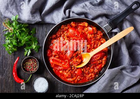 ragout with whole plum tomatoes, onion slices, ground italian pork sausages and spices in a skillet on a dark oak wooden table with grey cloth, flat l Stock Photo