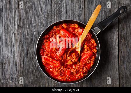ragout with whole plum tomatoes, onion slices, ground italian pork sausages and spices in a skillet on a dark oak wooden table, view from above, flat Stock Photo