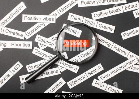 Magnifier glass over the red inscription marketing cut out of paper. Surrounded by other inscriptions on a dark background. Word cloud concept. Stock Photo