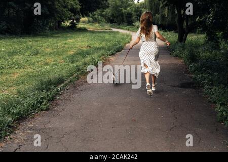 Young blonde woman running with a cat on a leash on the road in the summer park. Photo from the back of the general plan. The girl is dressed in a white long polka dot dress and white shoes. 