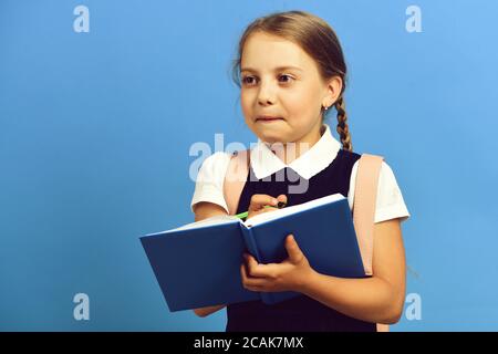 Girl writes in big blue notebook. Pupil in school uniform with braid and pink backpack. Back to school and education concept. School girl with scared face isolated on blue background Stock Photo