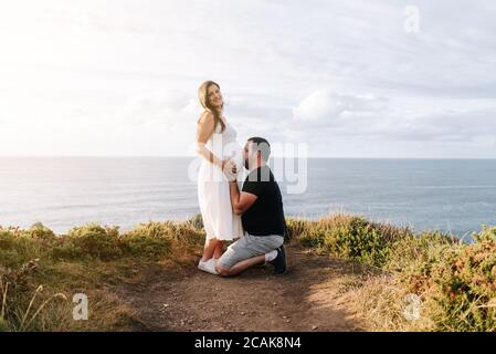 Pregnant couple with the father kissing the belly of his wife near the ocean Stock Photo