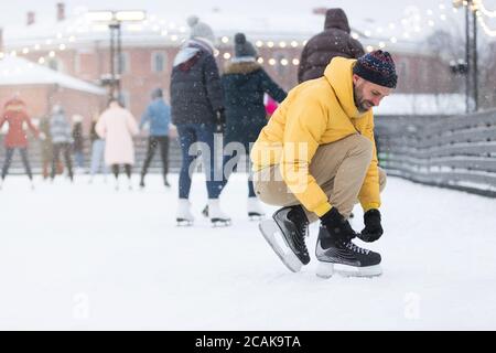 Portrait of Caucasian Scandinavian bearded man in yellow jacket, beige trousers wearing black skates on ice rink, side view.Caucasian young man tying Stock Photo