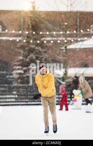 Portrait of joyful Scandinavian bearded man in yellow jacket, beige trousers on ice rink, outdoors in snowy winter day, Christmas tree on background. Stock Photo