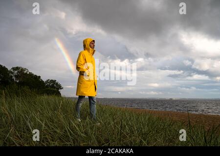 Man in yellow raincoat with hood walk on the beach in rainy weather, looks at dramatic cloudy sky and sea, rainbow on background. Autumn season. Stock Photo
