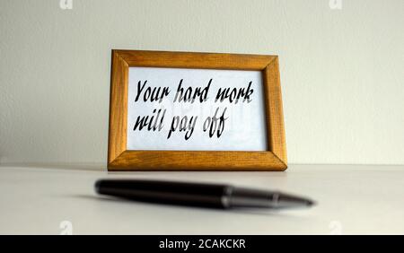 Wooden picture frame with inscription 'your hard work will pay off' on beautiful white background. Pen on the table. Stock Photo