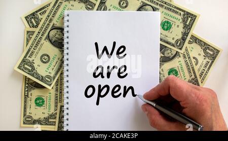 Male hand writing 'we are open' on white note, on white background. Dollar bills. Stock Photo