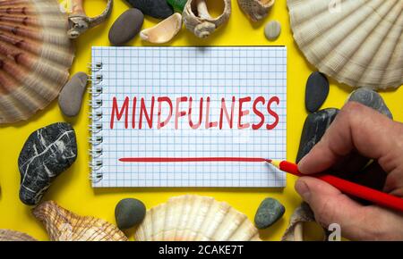 Male hand with red pencil writing 'mindfulness' on white note. Beautiful yellow background. Sea stones and seashells. Mindfulness concept, mindful liv Stock Photo