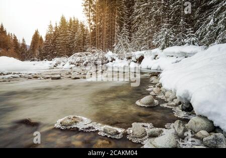 Morning sun shines on forest river creek in winter, rocks on shore covered with snow, long exposure makes water silky smooth
