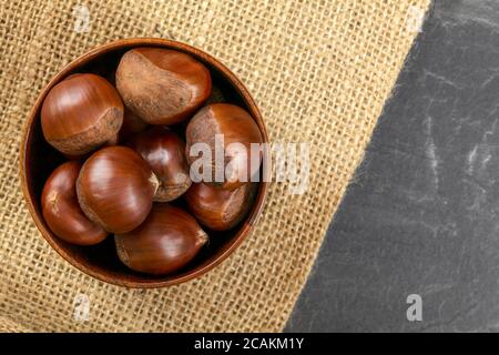 Tabletop view, small wooden bowl with chestnuts on jute cloth and slate like table Stock Photo