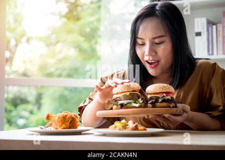 Hungry overweight woman holding hamburger on wooden plate after delivery man delivers foods at home. Concept of binge eating disorder (BED) and Relaxi Stock Photo