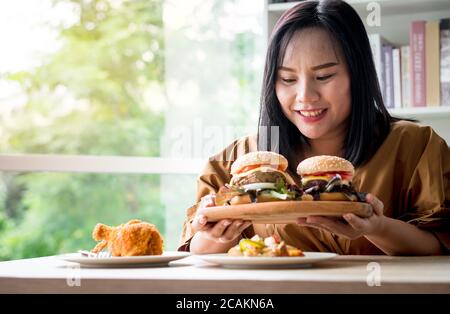 Hungry overweight woman holding hamburger on wooden plate after delivery man delivers foods at home. Concept of binge eating disorder (BED) and Relaxi Stock Photo