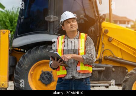 Engineering wearing a white safety helmet standing in front of the backhoe Looking at home construction work And use the tablet to check the blueprint Stock Photo