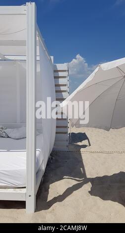 Geometry of white sunshade and beach umbrella shade on clear beach sand. Striped wooden wall painted in white color with blue sky and cumulus clouds. Stock Photo