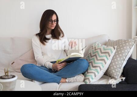Young woman sitting on sofa and reading book. Woman alone at her home reading a book with a cup in hand. She is comfortably sitting in the armchair. Stock Photo