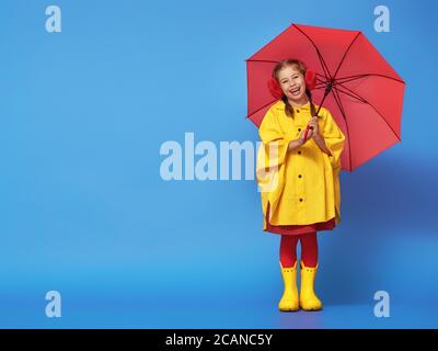 Happy funny child with red umbrella posing on blue wall background. Girl is wearing yellow raincoat and rubber boots. Stock Photo