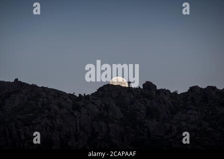 Almost full moon slowly rises behind stones on mountain peaks in a twilight moment, far away in the distance Stock Photo