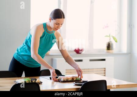 Young woman waiting for guests, friends while arranging the table with various sushi sets in a sunny home with window in the background Stock Photo