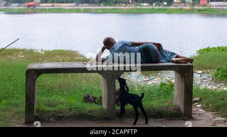 A man is resting on the bank of a river and his goats are eating grass Stock Photo