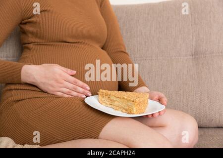 Close up of hungry pregnant woman is eating a piece of tasty cake relaxing on the sofa at home. Sweet cravings during pregnancy. Stock Photo
