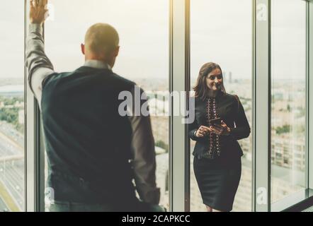 Silhouettes of two entrepreneurs near the window with selective focus on businesswoman with a smartphone in hands, her partner a businessman Stock Photo
