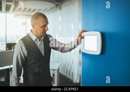 A mature handsome caucasian man entrepreneur using a multifunctional wall-mounted terminal panel with a blank white screen mock-up to adjust light Stock Photo