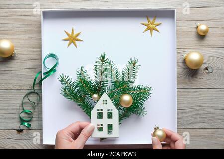 Christmas decorations, flat lay with hands making decorated box with house, fir twigs and golden trinkets, baubles. Flat lay on wooden table. Stock Photo