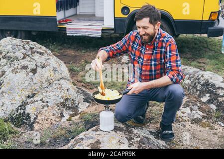 Young man cooking in campervan while staying in the countryside. Self built off-grid motorhome camping in the wild nature. Stock Photo