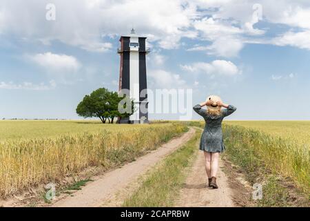 Woman in straw hat walking through the rye field towards lighthouse on summer day Stock Photo