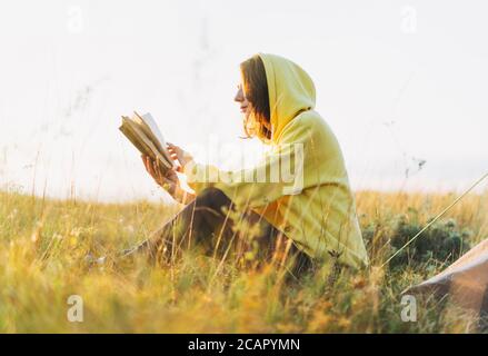 Young woman girl traveler in yellow hoodie reading book in morning sunrise. Local tourism, weekend trip Stock Photo