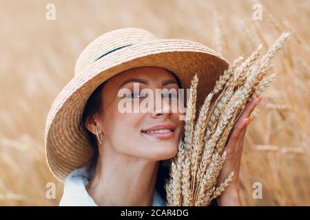 Young Woman in straw hat holding sheaf of wheat ears at agricultural field Stock Photo