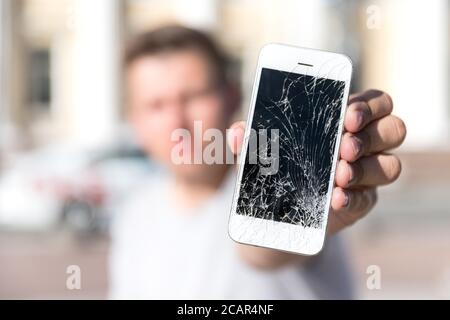 Caucasian young male holding and showing broken smartphone screen (display), selective focus on smartphone, blurred face, outdoors. Stock Photo