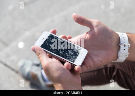 Male hands holding broken mobile smartphone after drop, view from above, outdoors. Smart phone with broken (cracked) screen. Picking broken phone from Stock Photo